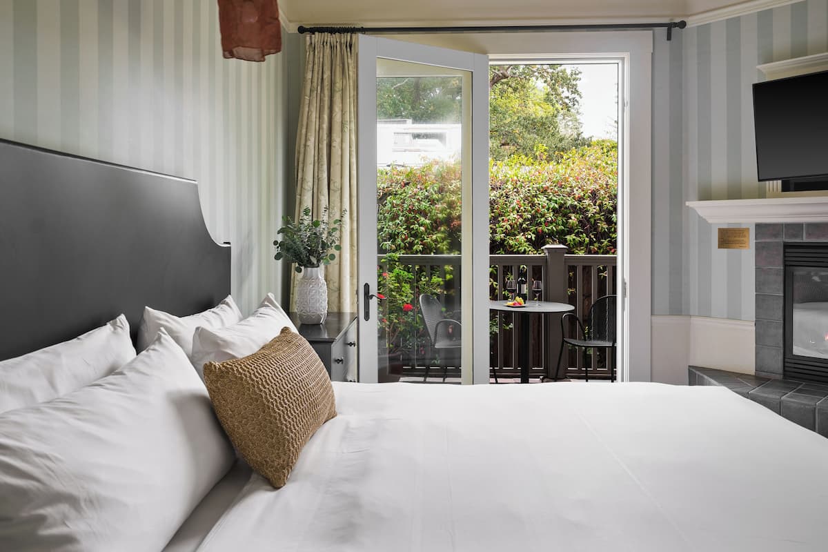 A hotel room with a large bed, a small table with chairs, a fireplace, and a view of a garden through a glass door.