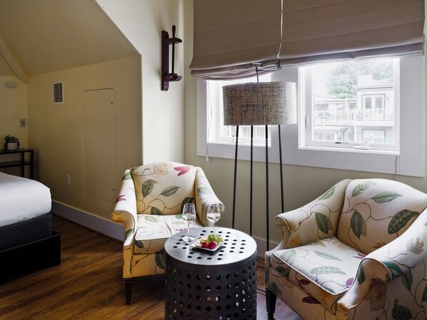 A cozy room with two floral-patterned armchairs, a small round table with a drink and snacks, a bed, and a window with a view of buildings. - Inn At Sonoma - A Four Sisters Inn (Sonoma)