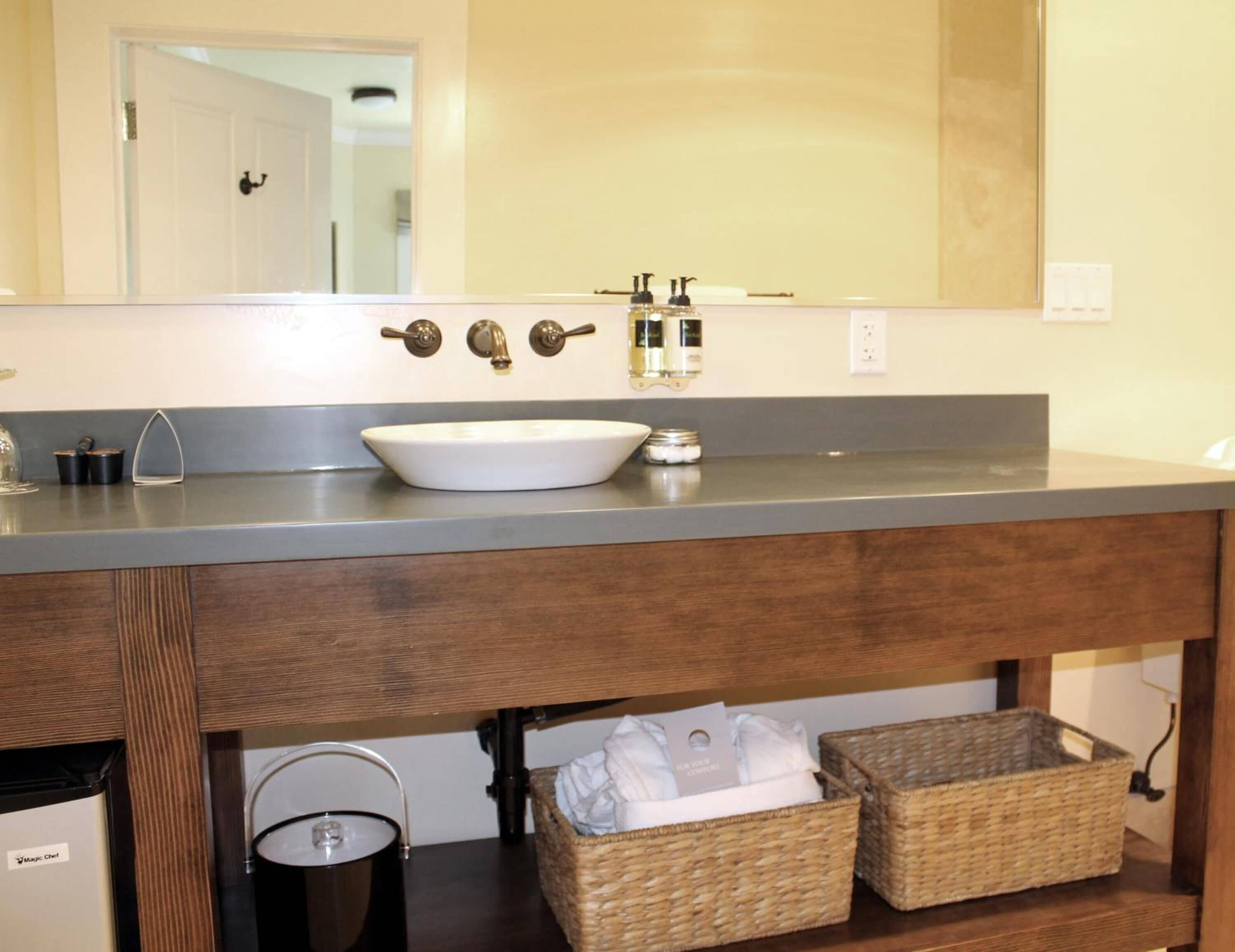 A bathroom with a white sink, two faucets, and a mirror above. There are wicker baskets under the sink and a trash can to the left.