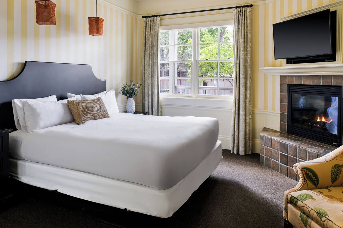 A hotel room with a large bed, a television mounted above a fireplace, and a window with curtains.