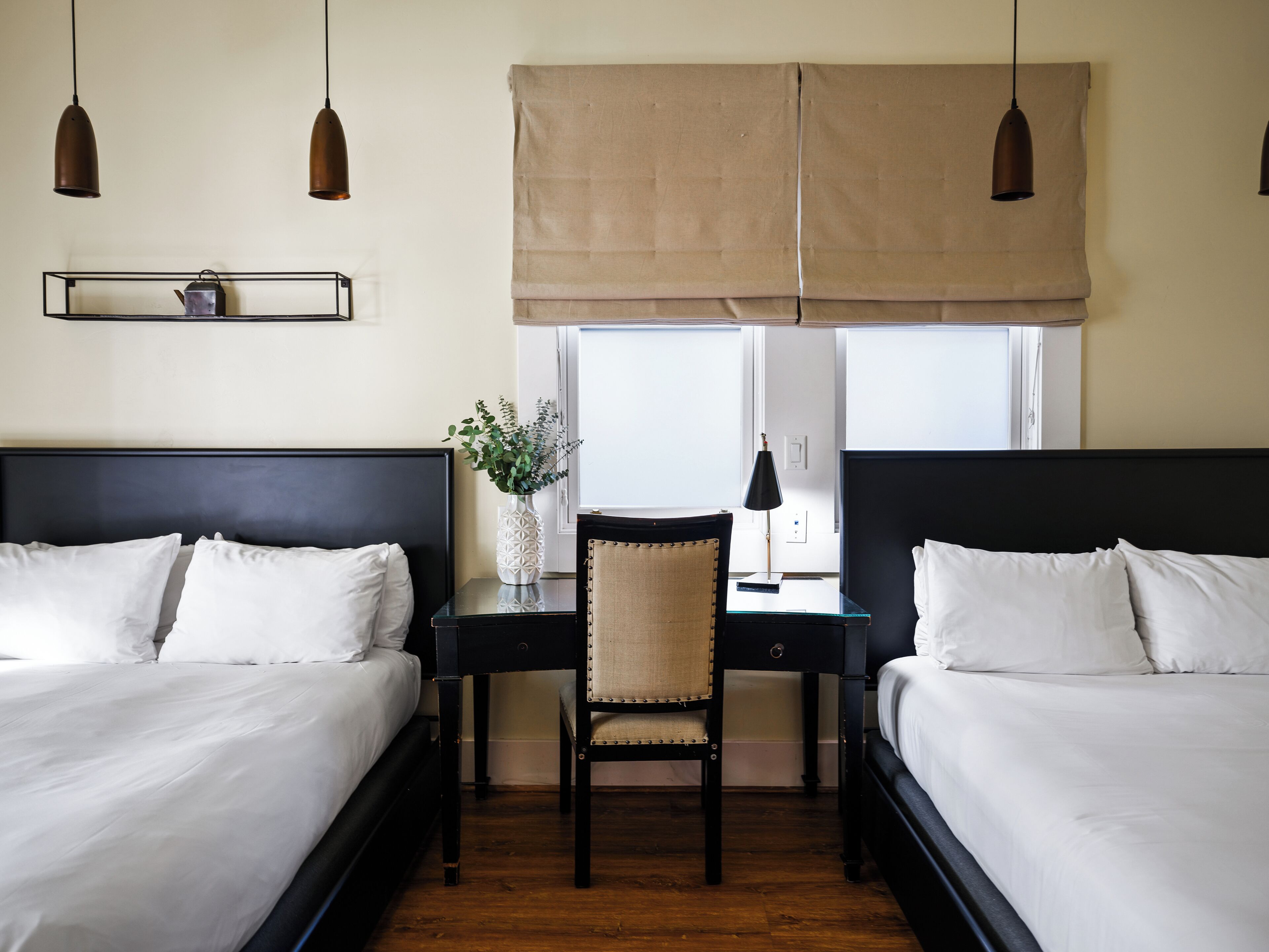 A hotel room with two beds, a desk, a chair, and a window with a beige blind.