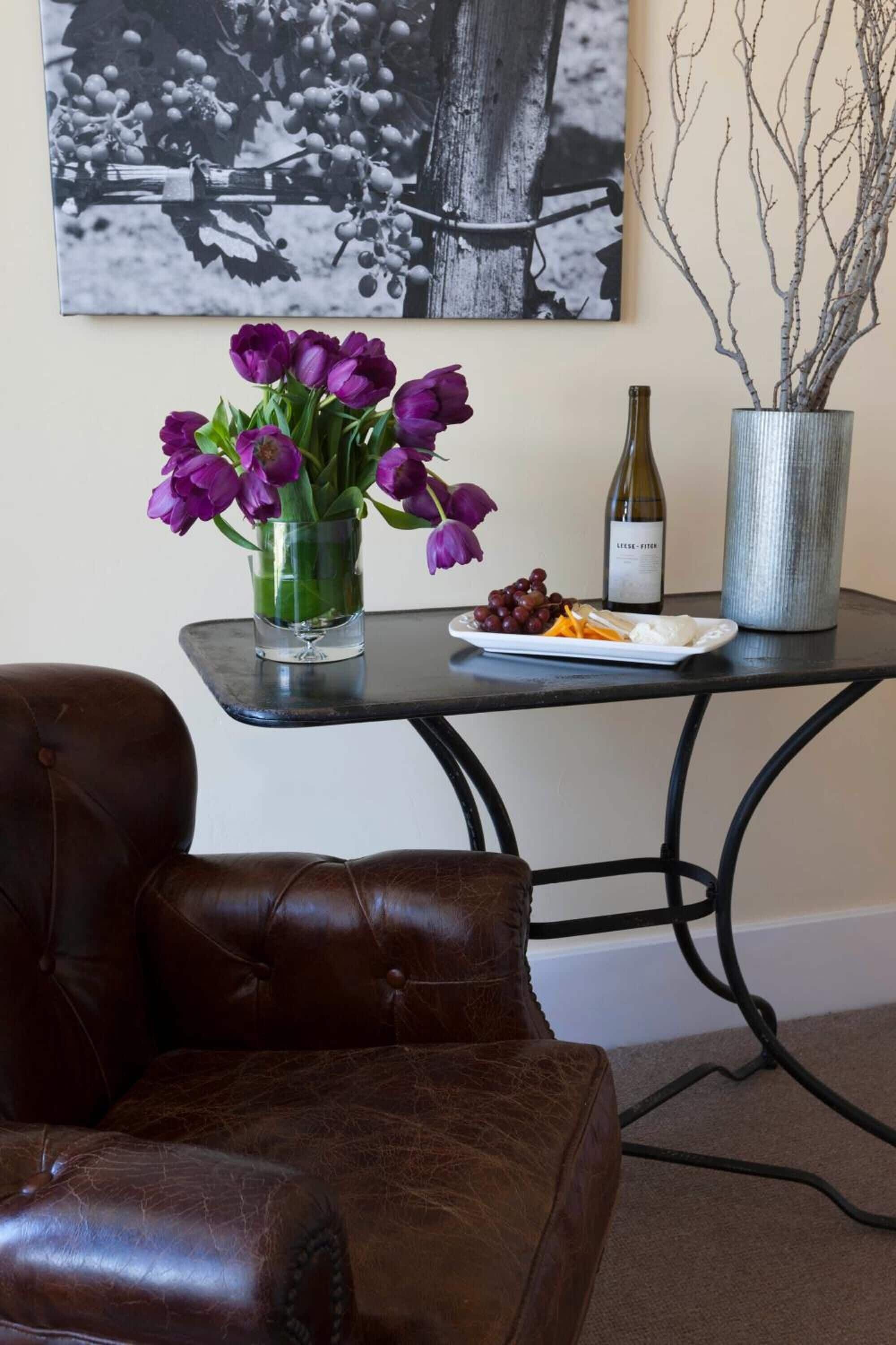 A brown leather chair, a small table with a vase of purple flowers, a plate of grapes, a bottle of wine, and a metal vase with branches.