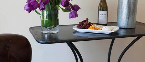 A brown leather chair, a small table with a vase of purple flowers, a plate of grapes, a bottle of wine, and a metal vase with branches.