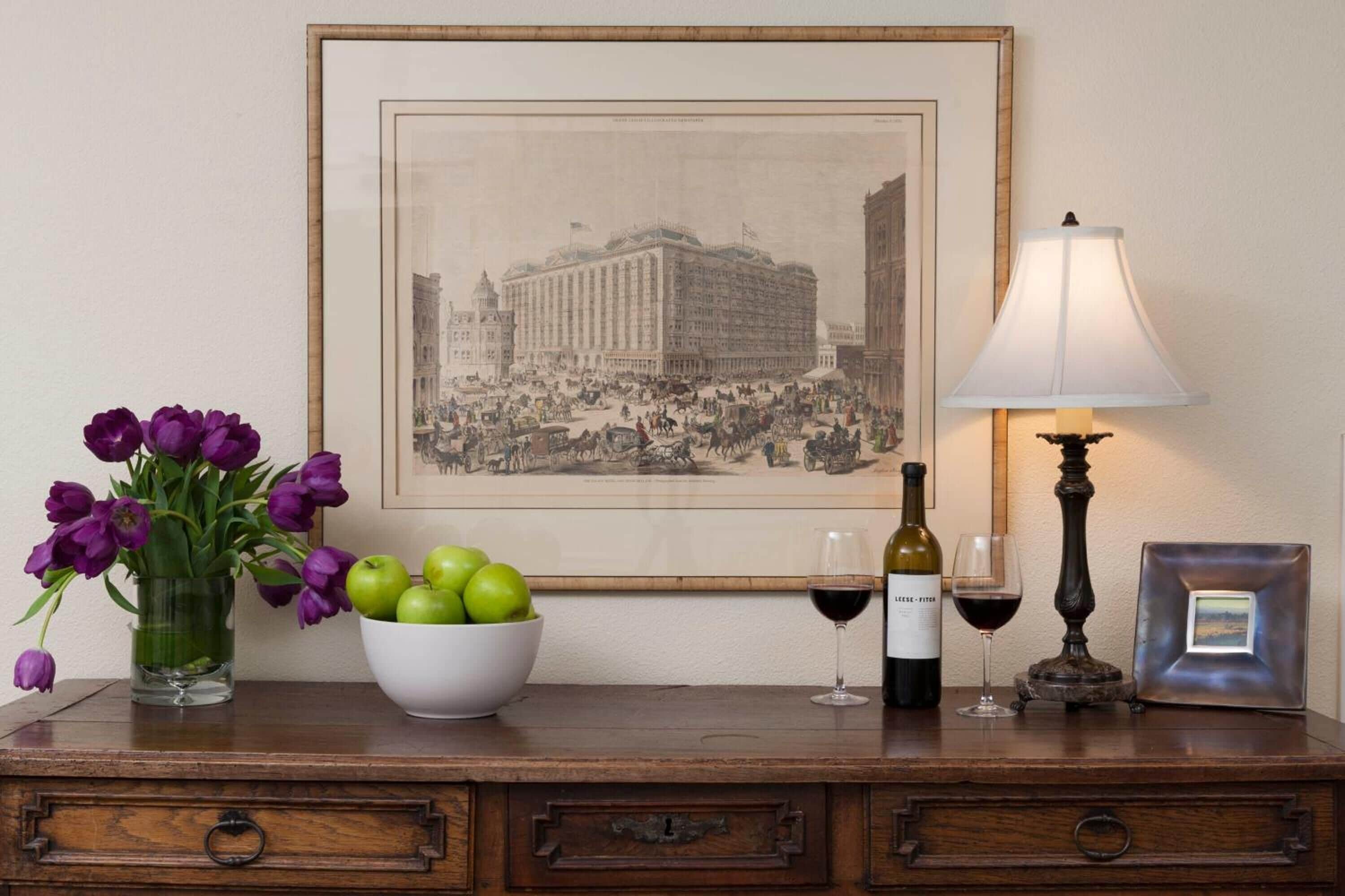 A wooden table with a framed black and white photograph, a vase of purple tulips, a bowl of green apples, a wine bottle, and a wine glass.
