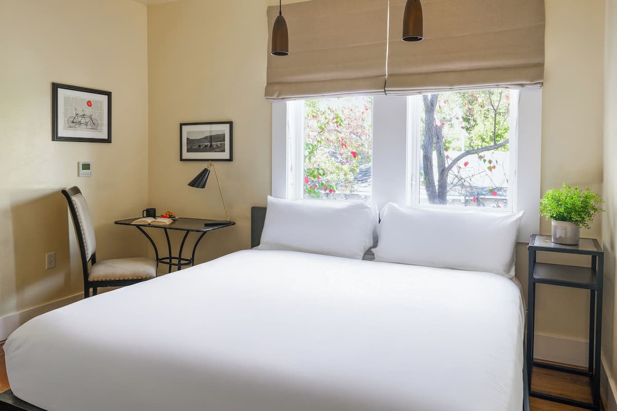 A neatly made bed with white linens, a small table with a lamp, a chair, and two framed pictures on the wall.