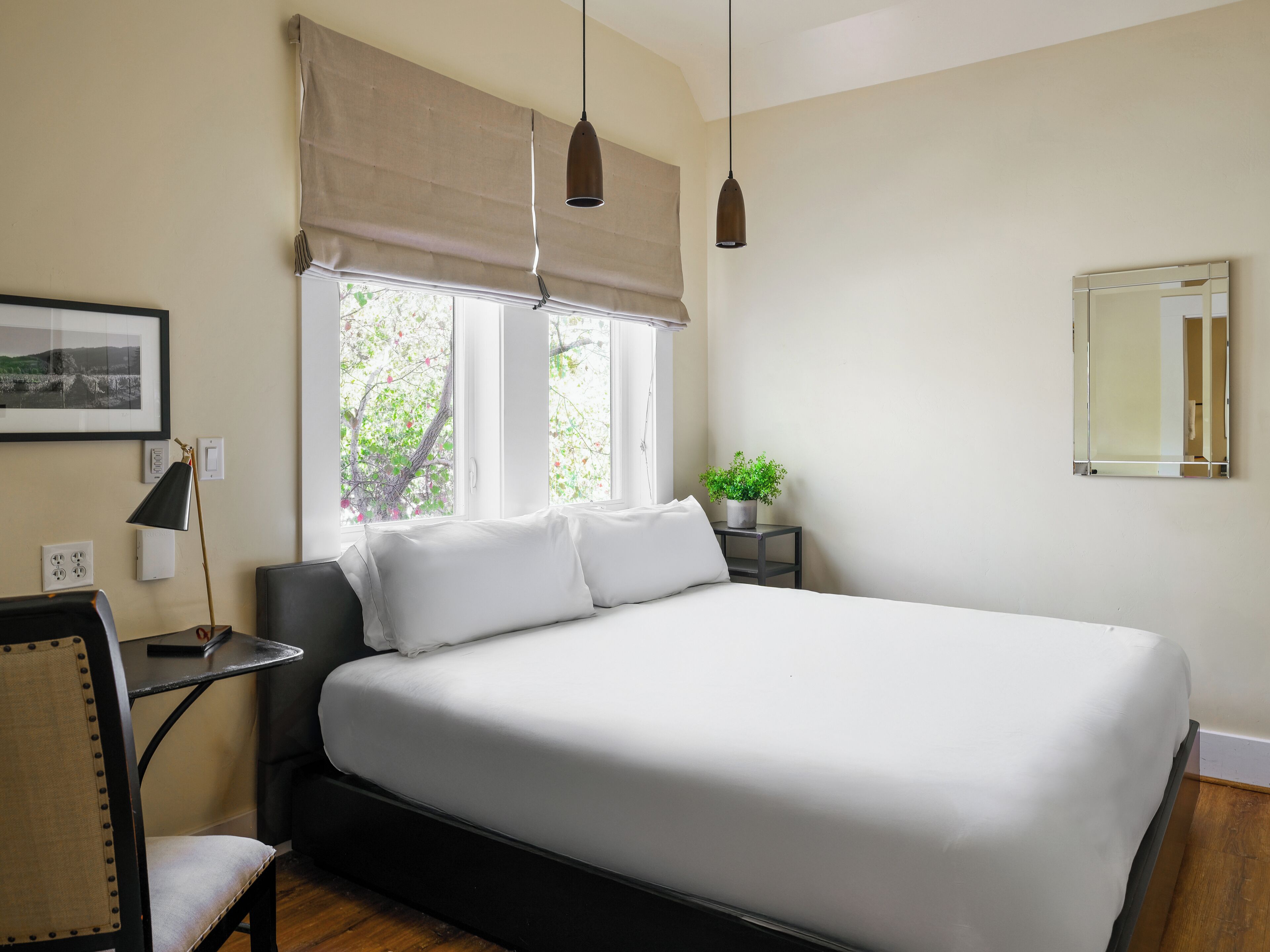 A bedroom with a bed, bedside tables, a chair, a lamp, a potted plant, and a window with a view of trees.