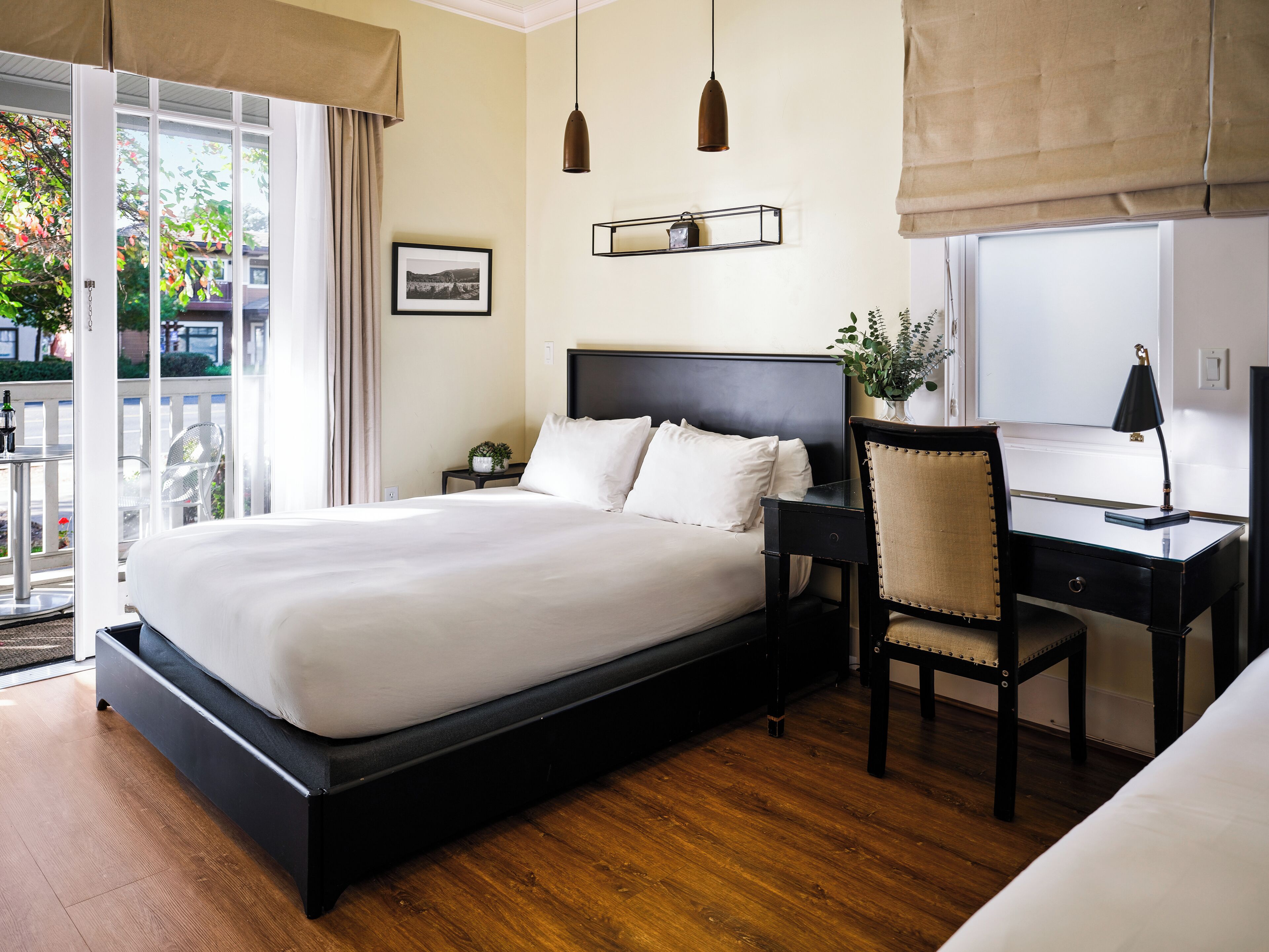 Quarto de hotel com uma cama grande, uma escrivaninha com cadeira, uma mesa de cabeceira, uma luminária pendente e um quadro na parede, além de uma janela com vista para o verde lá fora.