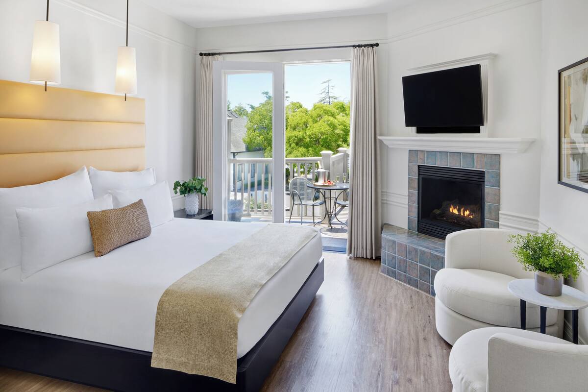 A modern hotel room with a large bed, a fireplace, a balcony with outdoor furniture, and a television mounted above the fireplace.