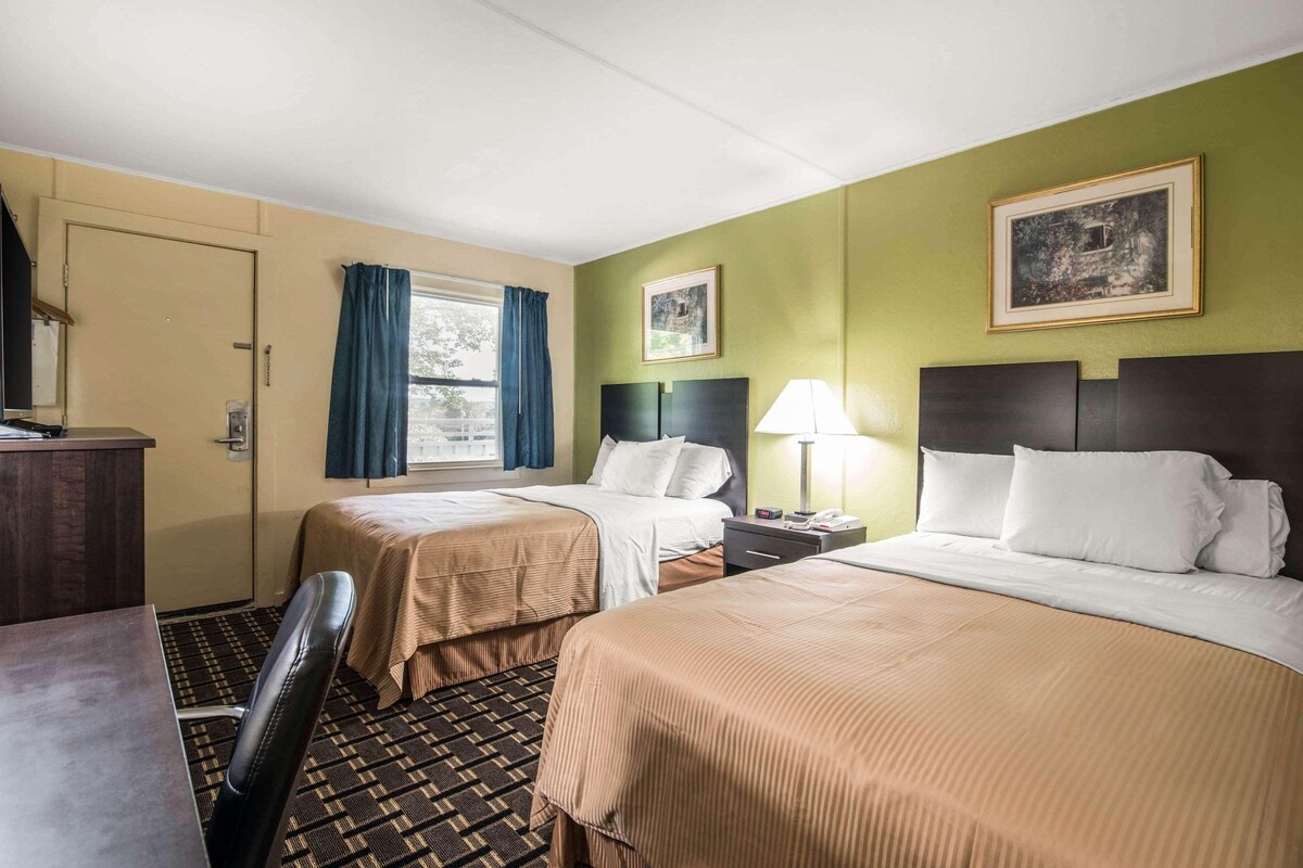 Una habitación de hotel con dos camas, un escritorio, una silla, un ventanal con cortinas y un cuadro en la pared.