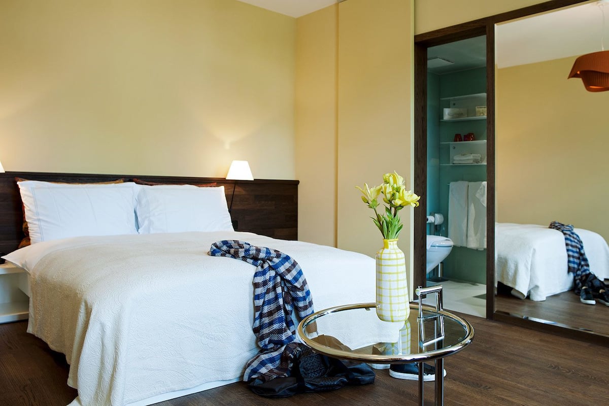 Habitación de hotel con cama, mesita de noche con un florero y un espejo que refleja el cuarto.