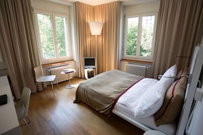 A hotel room with a bed, a desk, a chair, a TV, and large windows. - Design Hotel Plattenhof (Zürich)