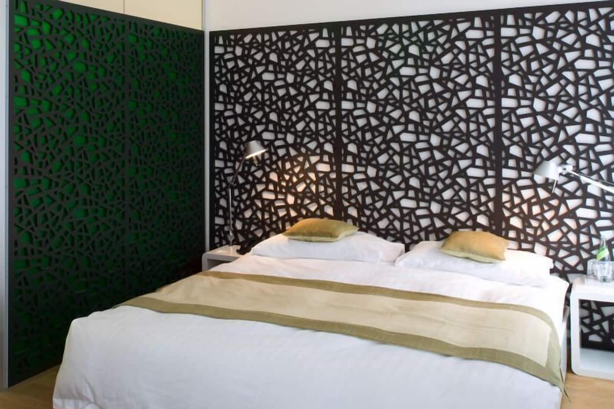 Una cama bien hecha con ropa de cama blanca y un detalle en color beige, frente a una pared con un diseño geométrico.