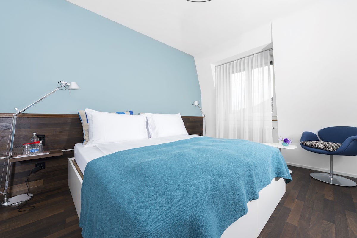 Habitación de hotel con una cama, una mesita de noche con lámpara, una silla y una ventana con cortinas.