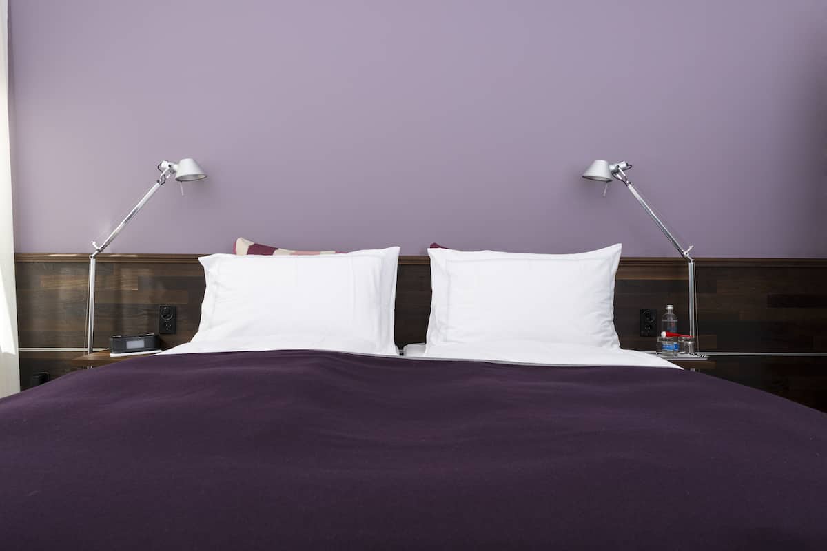 Una cama bien hecha con almohadas blancas y una manta púrpura, flanqueada por dos mesitas de noche con lámparas, en una habitación con pared morada.