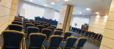 Un salón de conferencias con hileras de sillas azules y una larga mesa, piso alfombrado y una pizarra en el fondo.