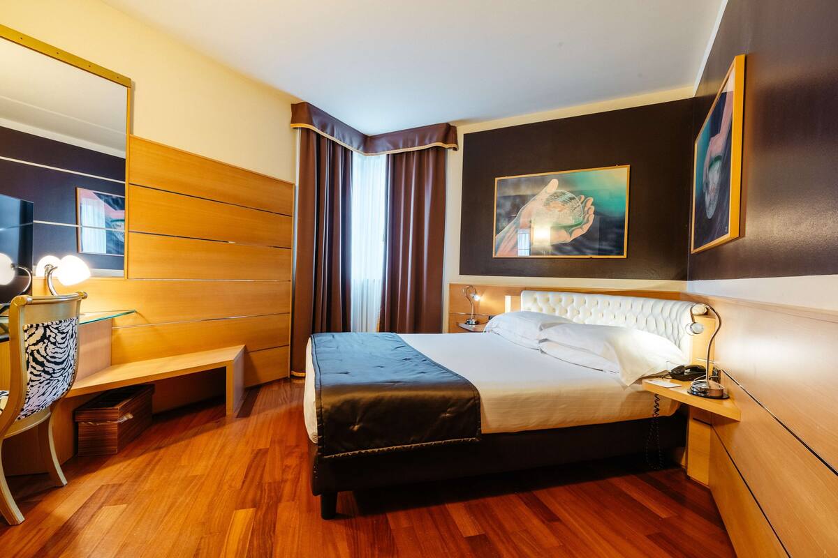 Una habitación de hotel moderna con una cama grande, mesita de noche, lámpara y un espejo.