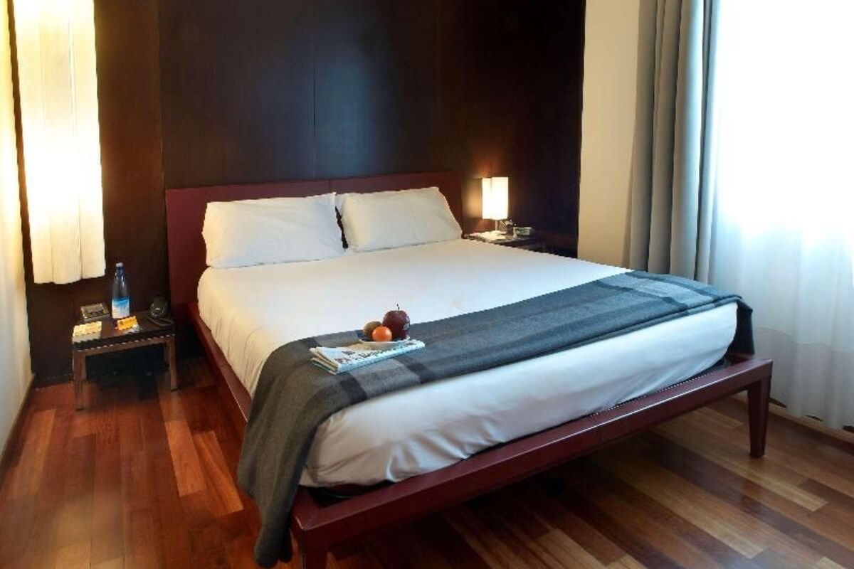 Une chambre d’hôtel avec un grand lit, un plancher en bois, une table de chevet avec une lampe et une fenêtre avec des rideaux.
