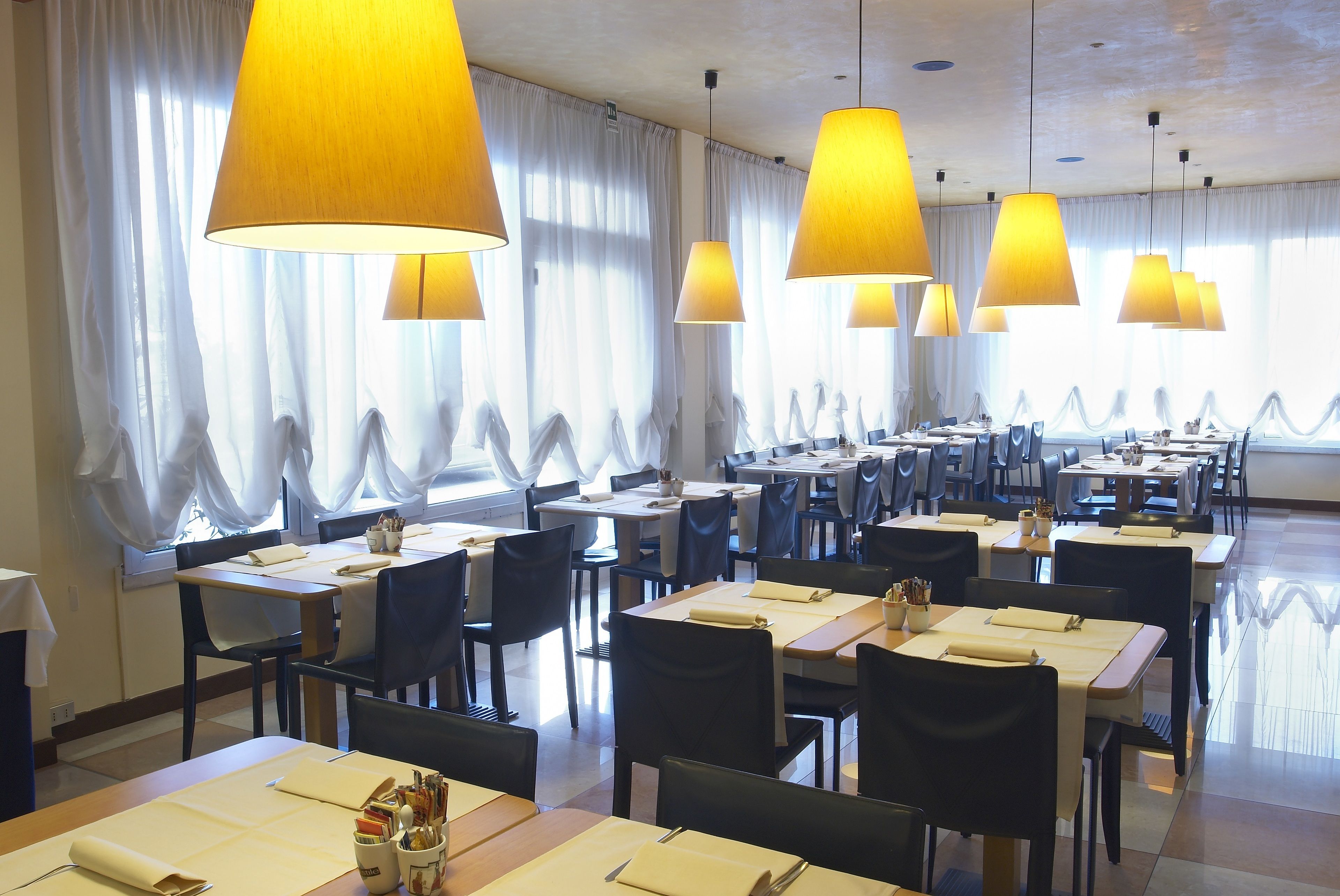 Un restaurante con varias mesas y sillas, grandes lámparas colgantes y vistas al exterior a través de amplios ventanales con cortinas traslúcidas.