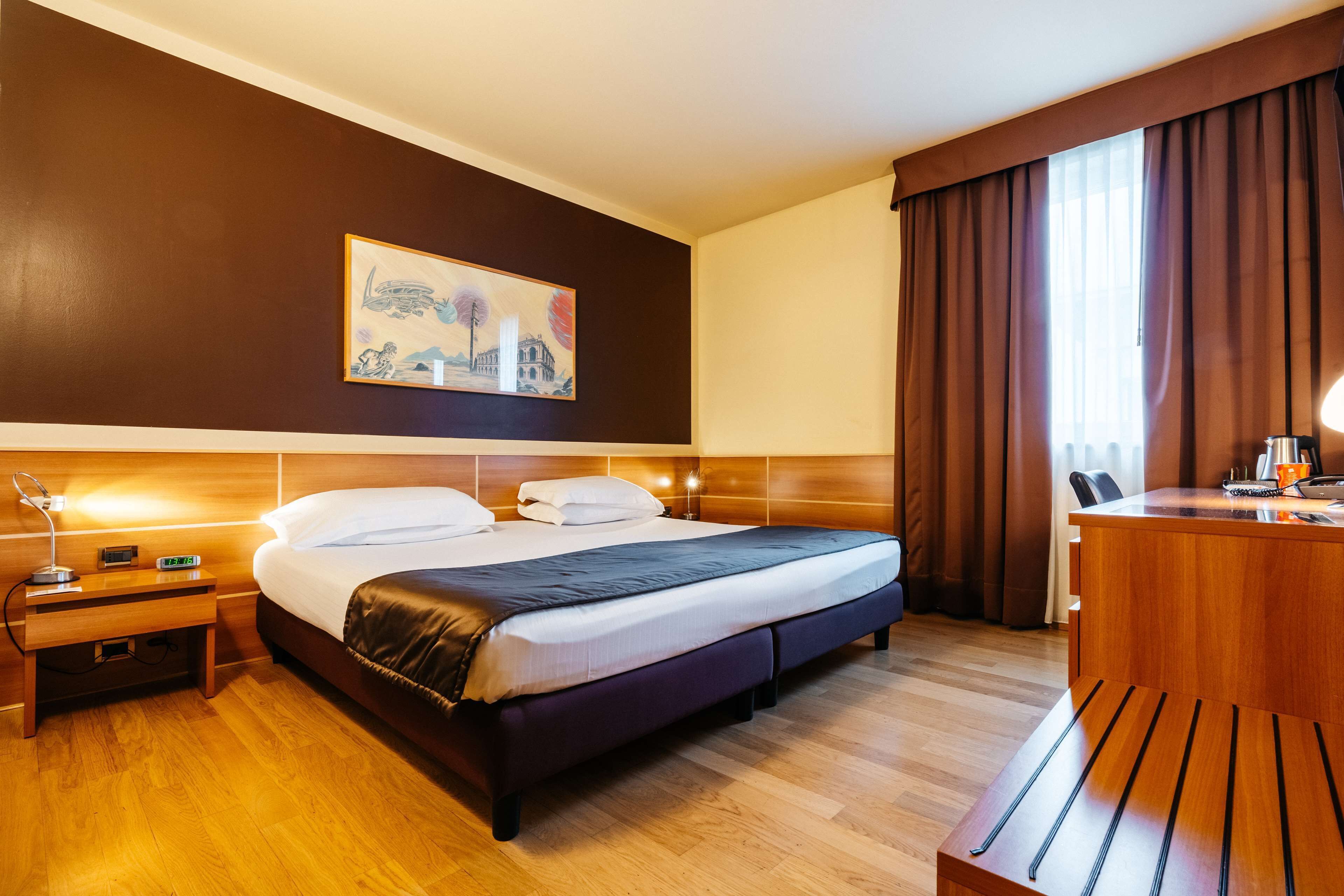 A hotel room with a bed, bedside table, lamp, and a painting on the wall.