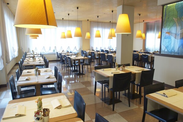 Un restaurant doté de nombreuses tables et chaises, de grands lustres suspendus et offrant une vue sur l’extérieur par les fenêtres.