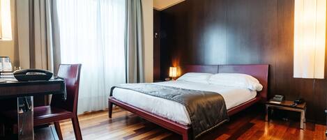 A hotel room with a wooden floor, a bed with a dark headboard, a desk with a chair, a lamp, and a window with curtains.
