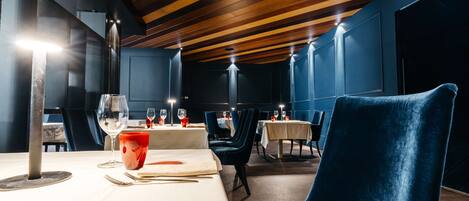 Un restaurante con vigas de madera en el techo, mesas dispuestas con manteles blancos y sillas azul oscuro.