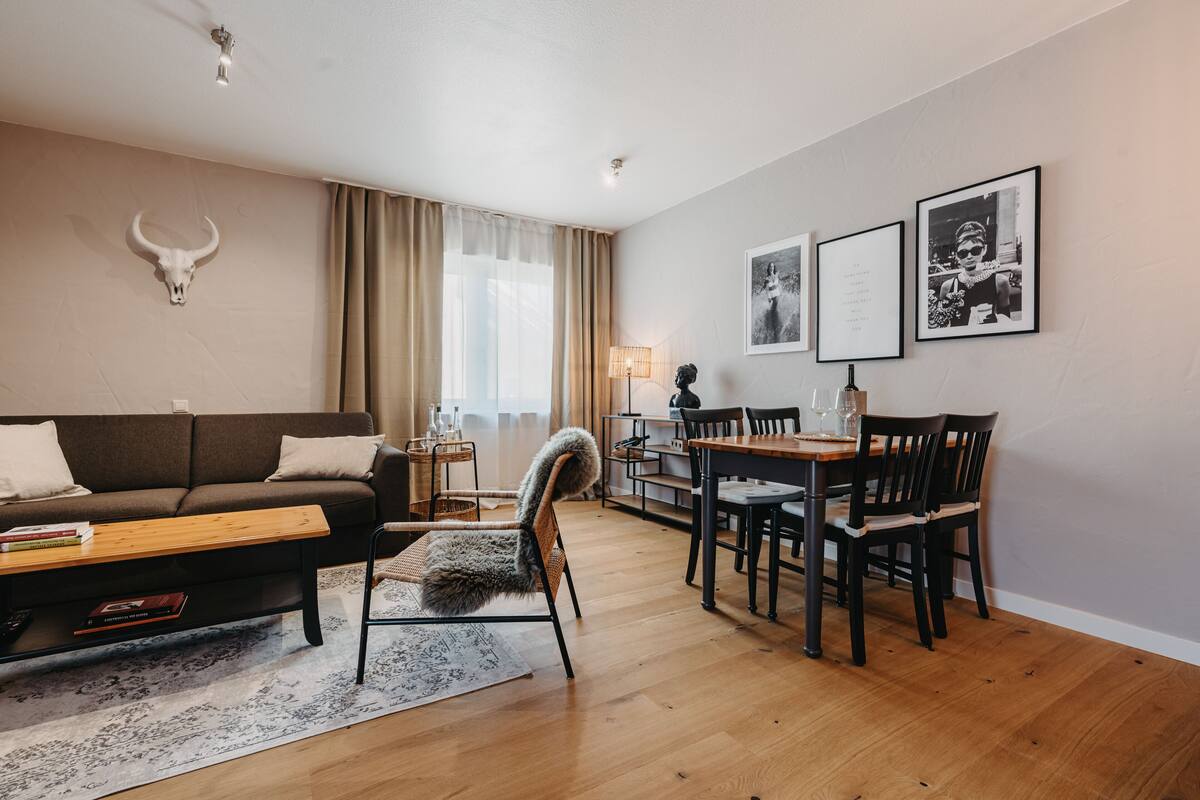 Uma sala de estar moderna com sofá, uma poltrona aconchegante, uma mesa de jantar com cadeiras, uma decoração de caveira de touro na parede e quadros emoldurados.
