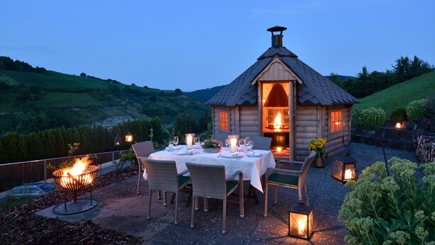 A cozy log cabin with a dining area set up outside, featuring a fire pit and lanterns. - MANA LISA Boutiquehotel (Lauda)