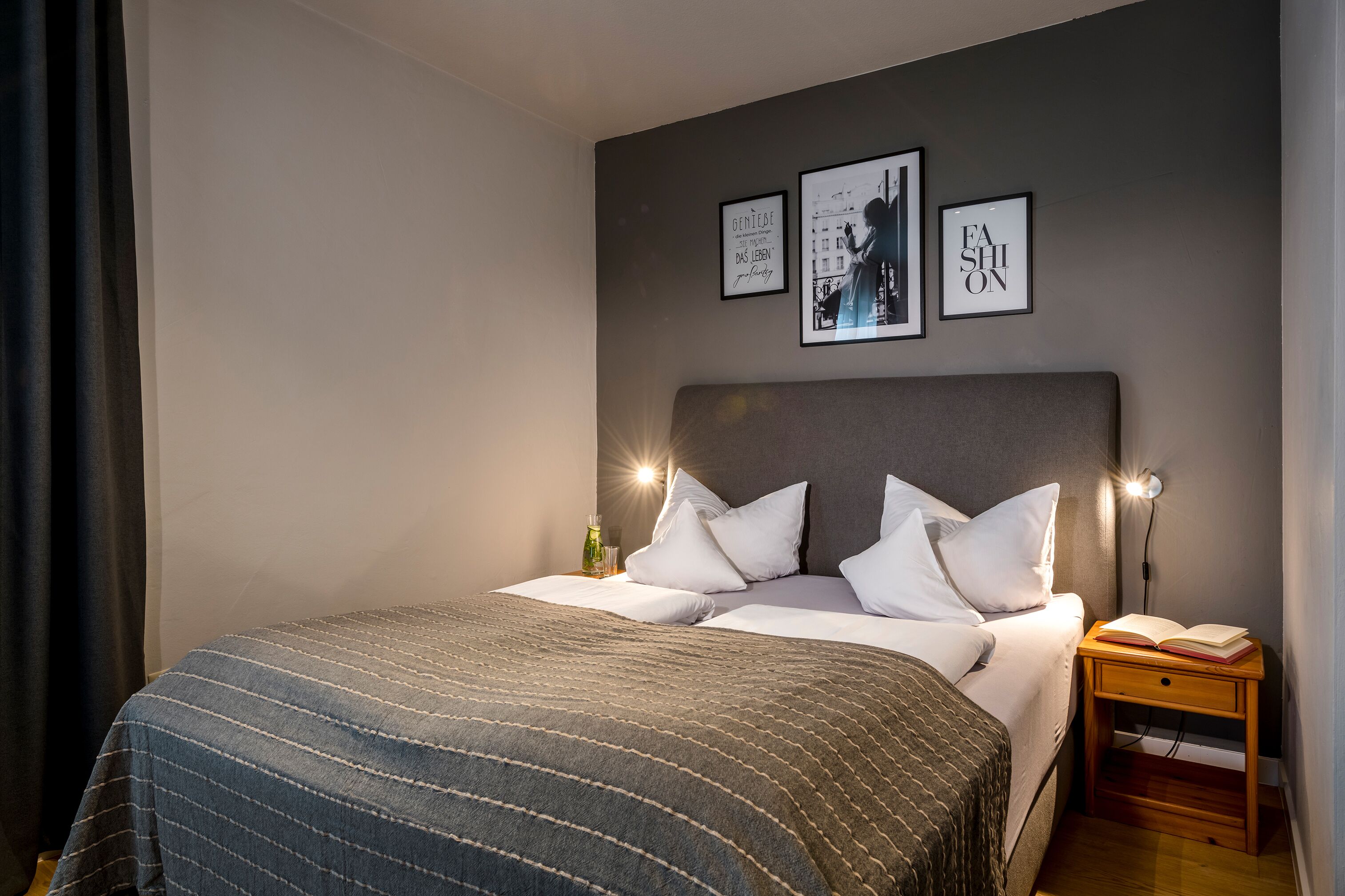 Una cama bien hecha con una manta a rayas, una mesita de noche con un libro y decoración en la pared que incluye una cita y una silueta.