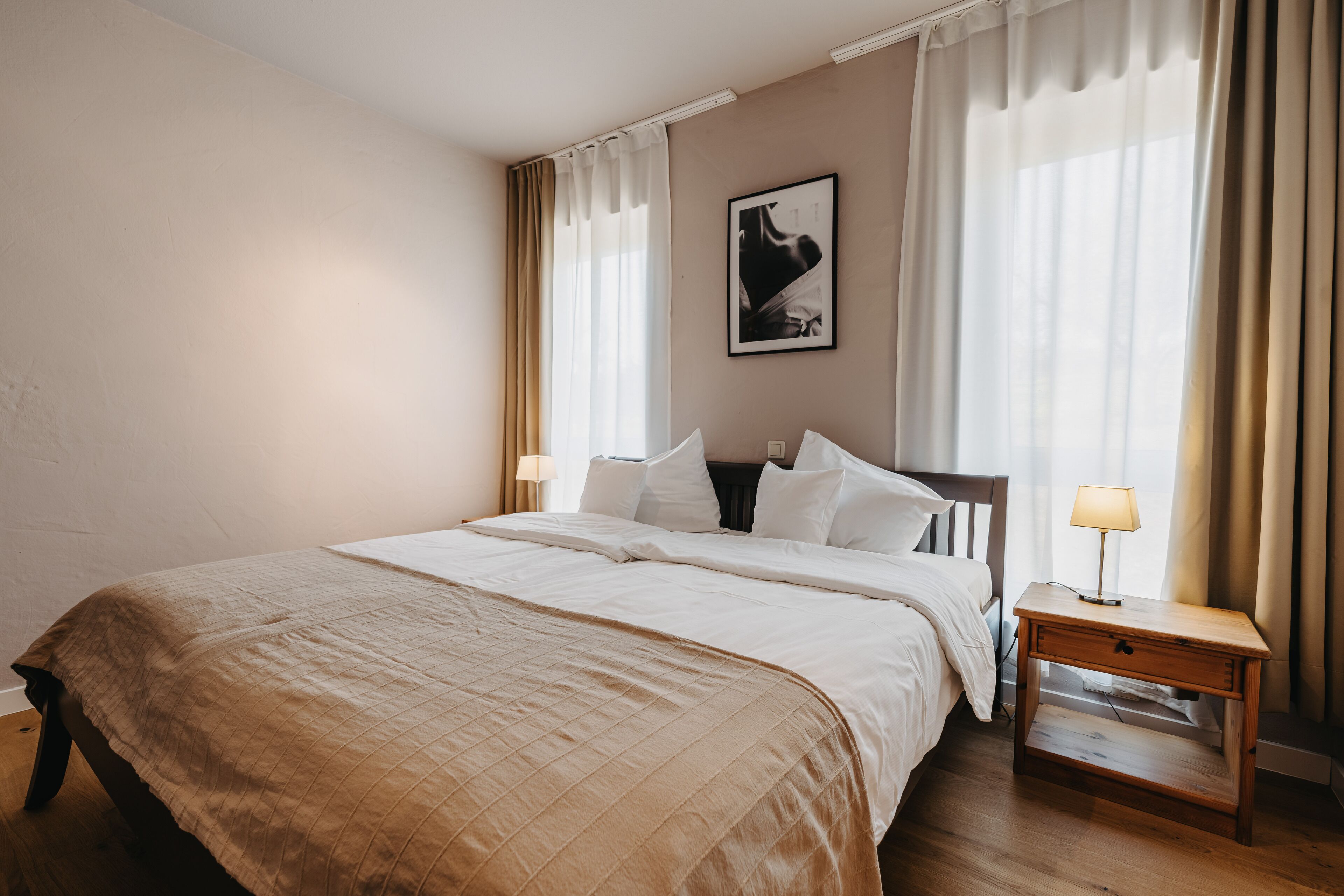 Una cama bien hecha con un edredón color beige, una mesita de noche con una lámpara y un cuadro en la pared.