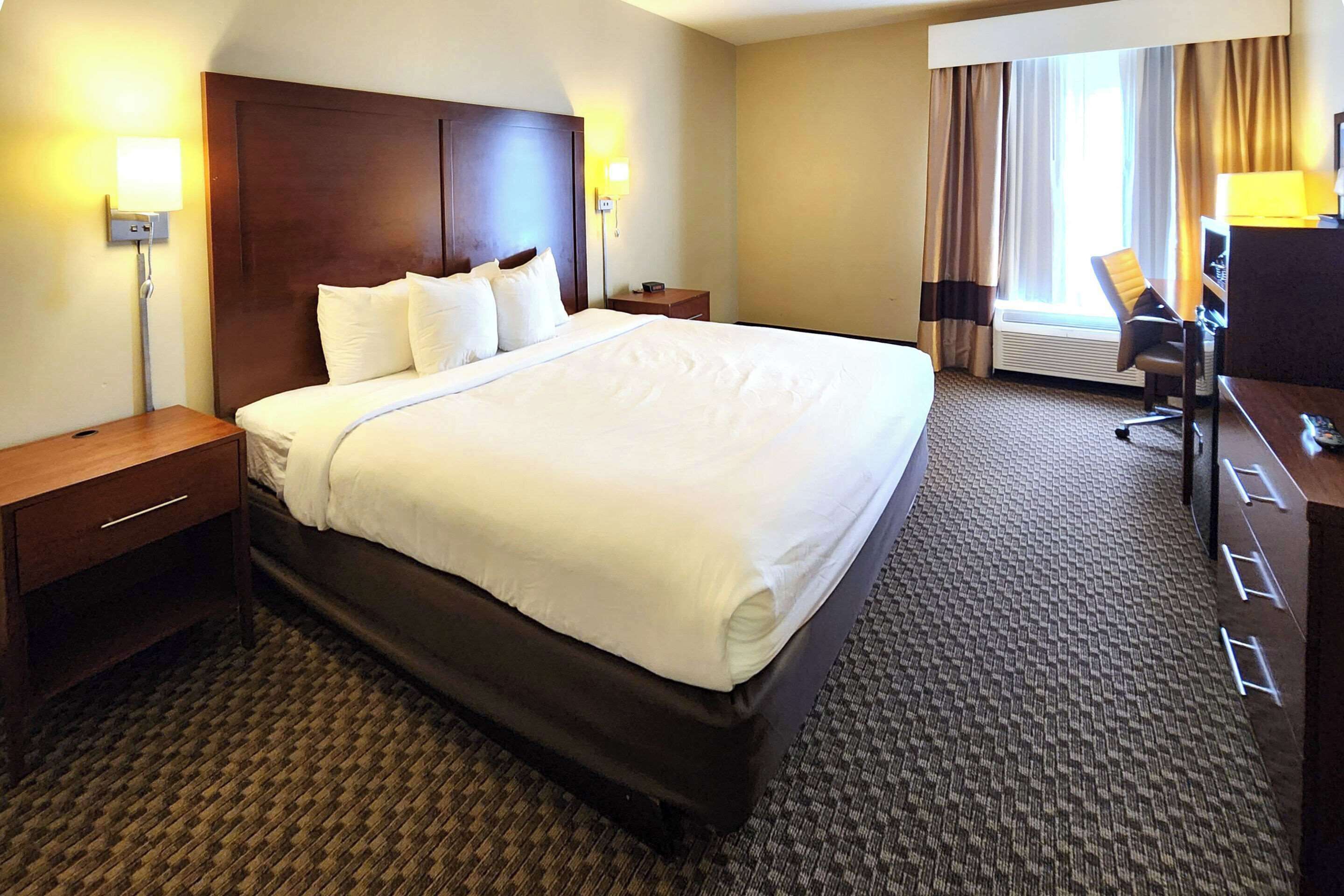 A hotel room with a large bed, two bedside tables, a desk with a chair, and a window with curtains.