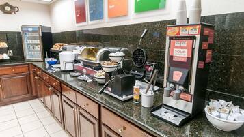 Una estación de desayuno con una máquina para hacer gofres, una cafetera y un bol de fruta.