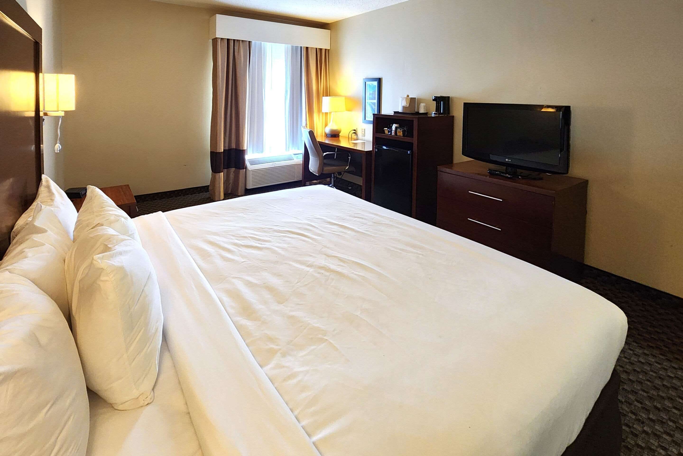 Quarto de hotel com uma cama grande, uma escrivaninha com cadeira, uma televisão e uma luminária de mesa.