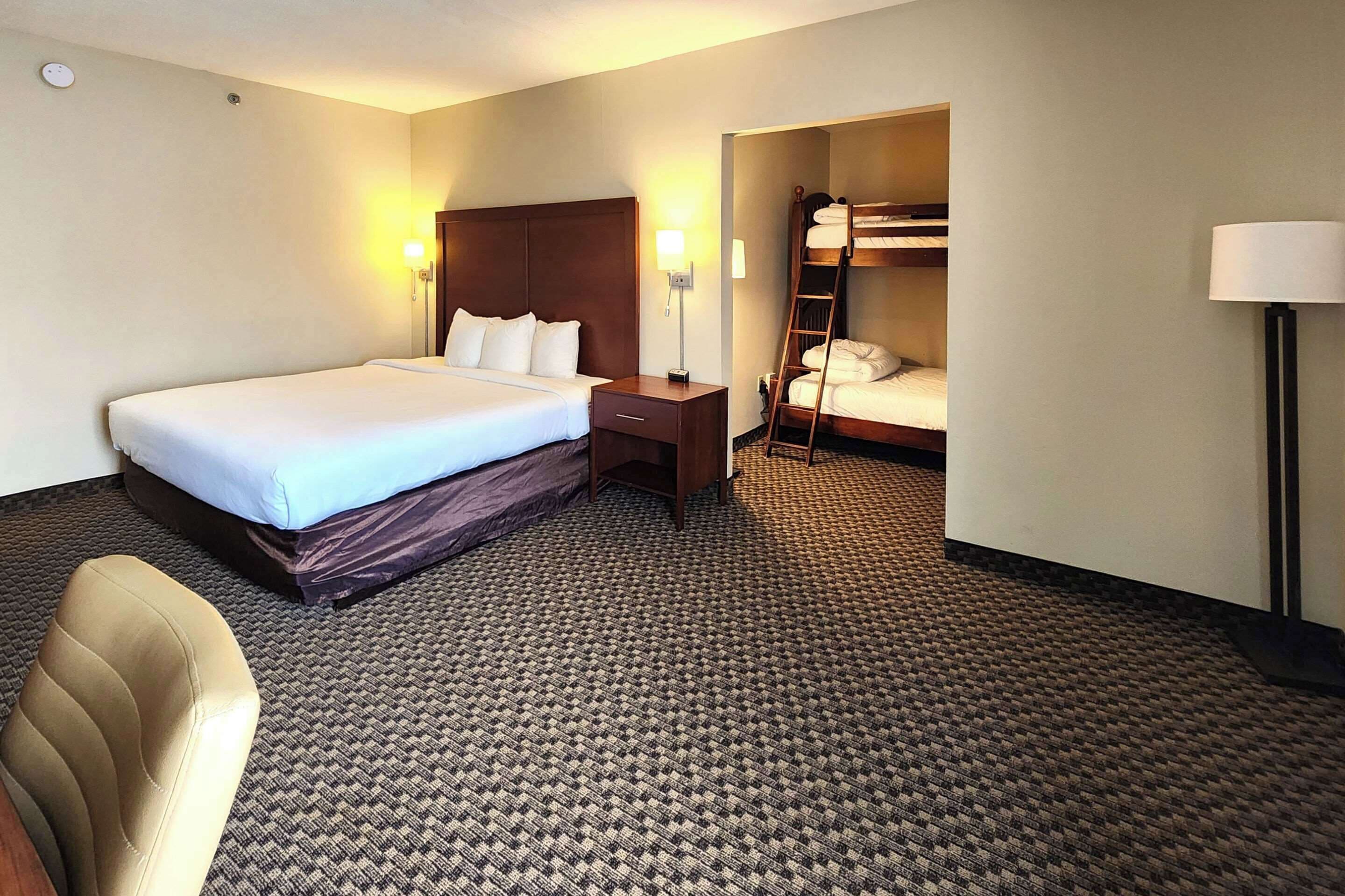 Habitación de hotel con una cama grande, un escritorio, una silla, una cama nido y una lámpara.