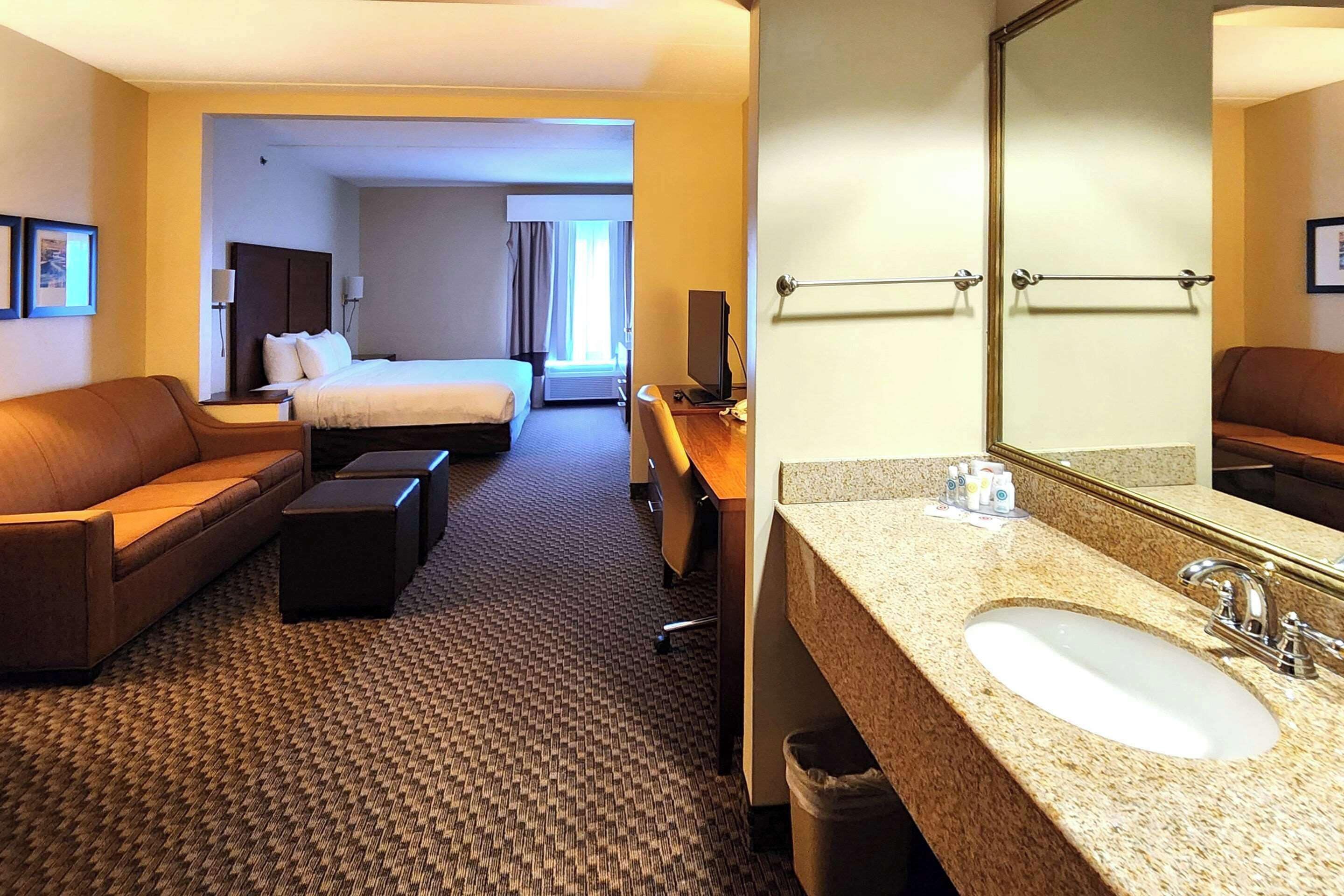 Habitación de hotel con cama, sofá, escritorio y baño con lavamanos y espejo.