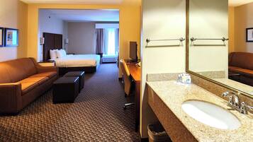 Habitación de hotel con cama, sofá, escritorio y baño con lavamanos y espejo.