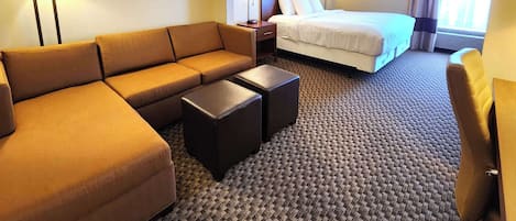 A hotel room with a sofa, two ottomans, a bed, a nightstand, and framed pictures on the wall.