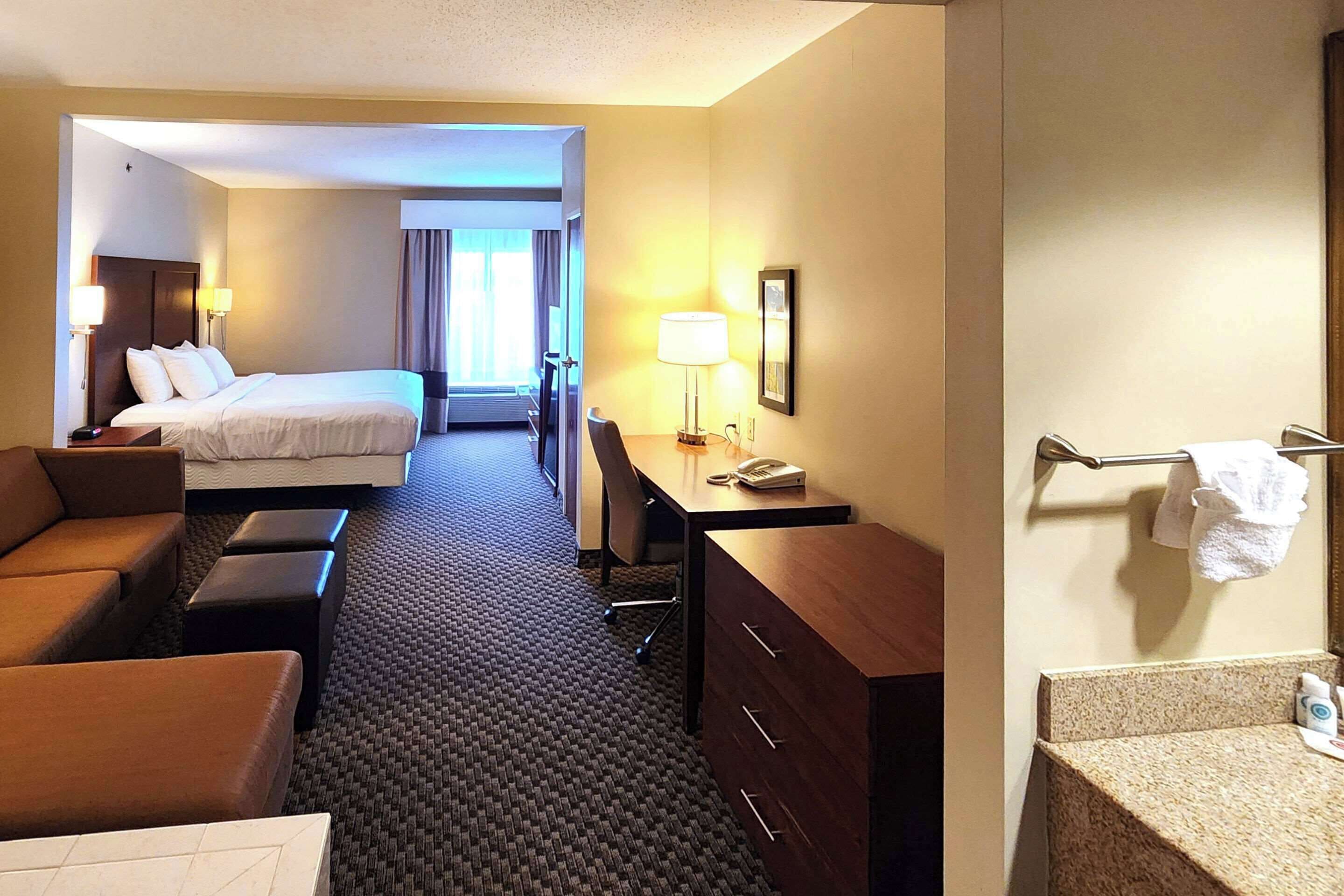 A hotel room with a bed, a desk, a chair, a sofa, a dresser, and a bathroom with a sink and mirror.