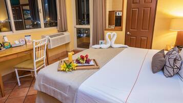 A hotel room with a neatly made bed, a breakfast tray with fruits and flowers, a desk with a chair, and a view of the city through the window.