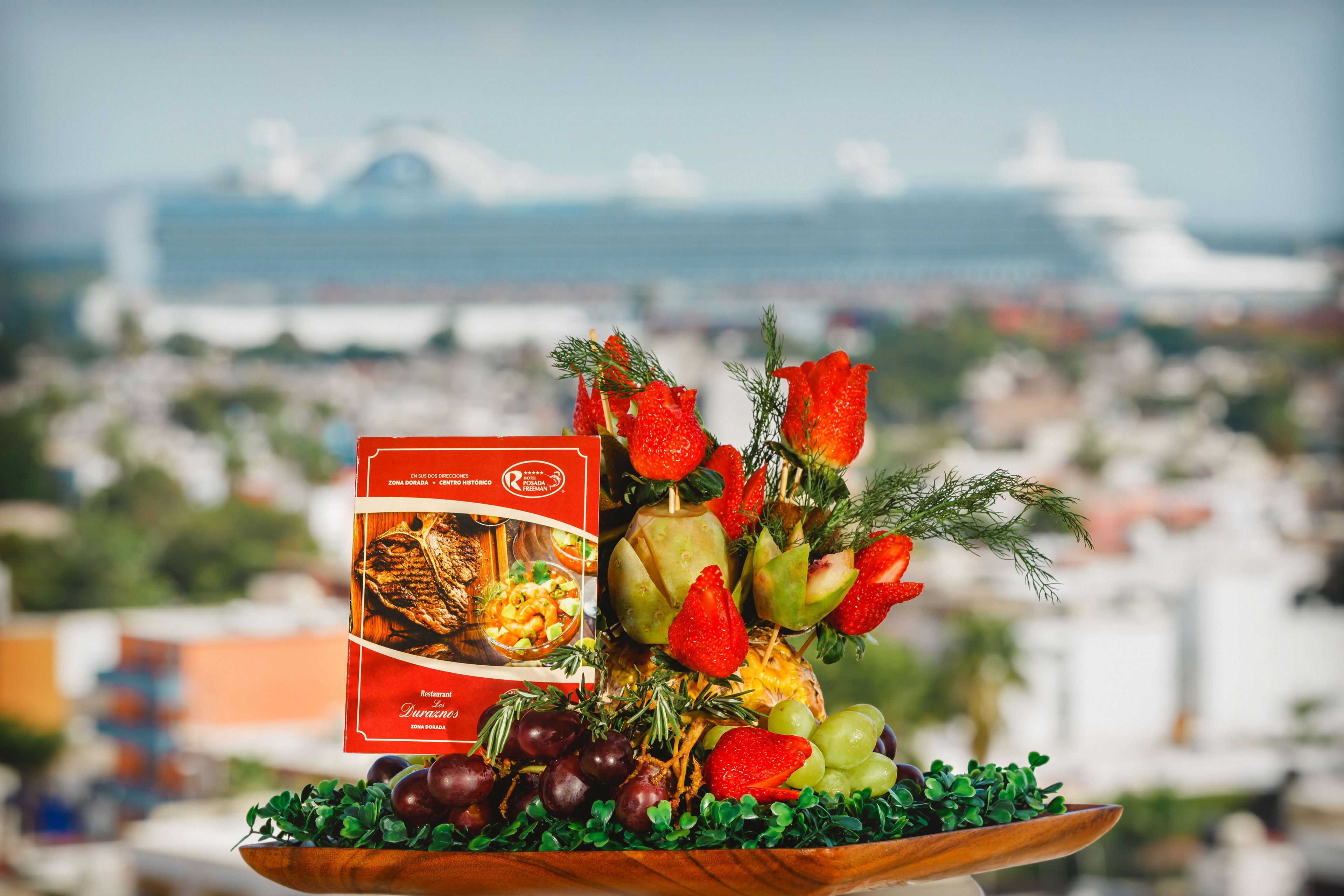 A fruit and cheese board with a brochure, set against a cityscape with a cruise ship in the background.