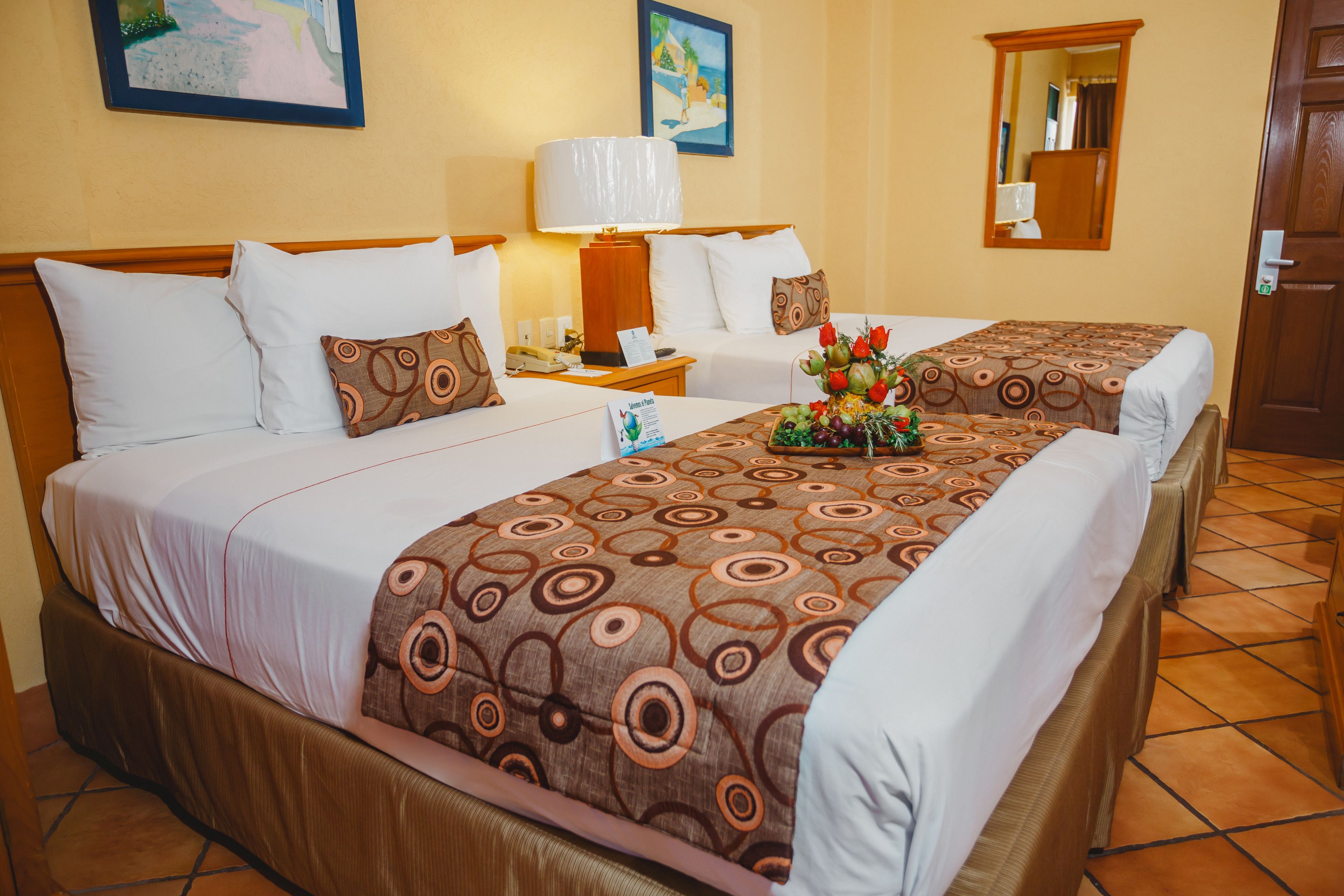 A hotel room with two beds, a nightstand with a lamp, a mirror, and a floral arrangement on the bed.