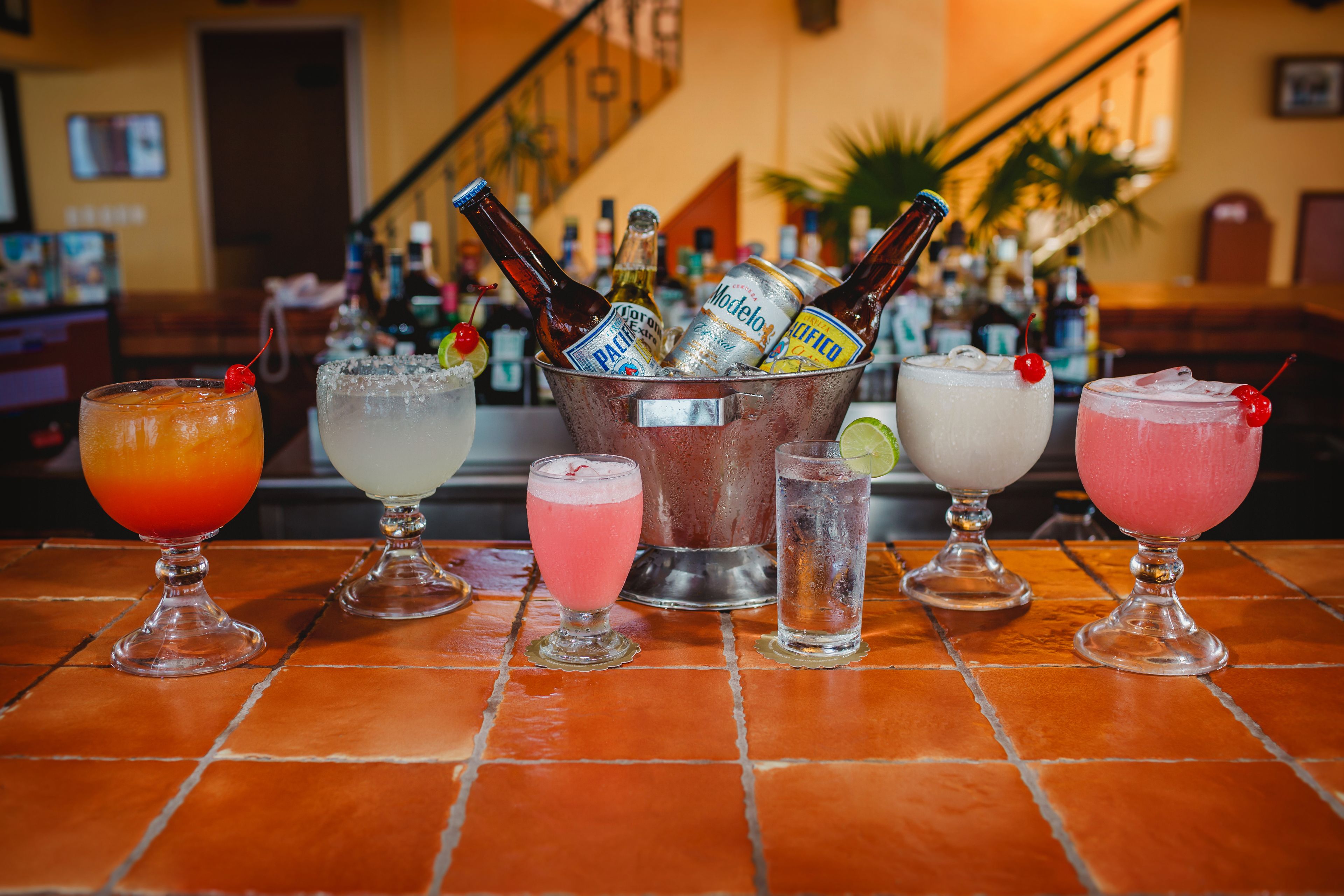 A bar with a variety of drinks in glasses and bottles, a tiled floor, and a background with a staircase and bar shelves.