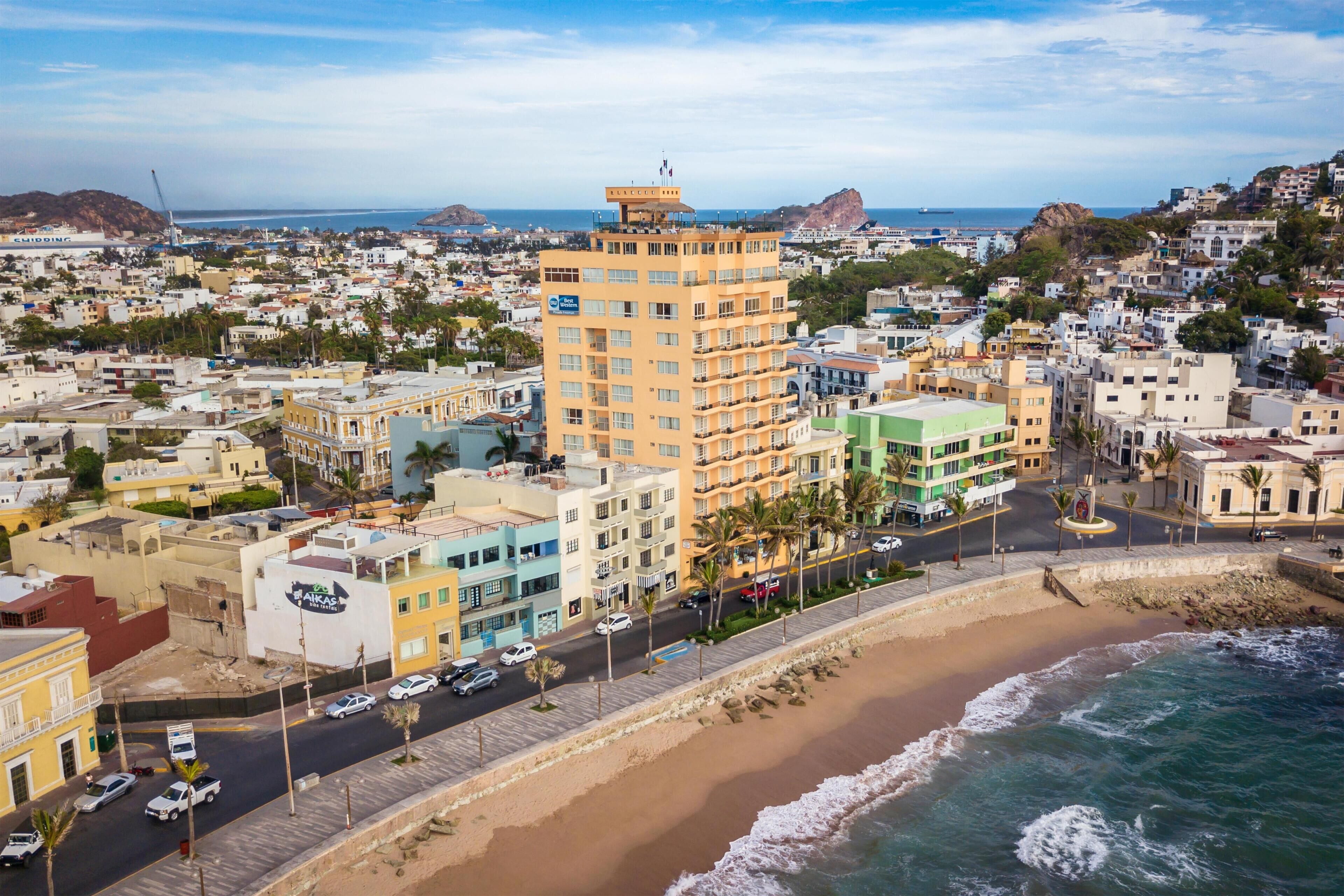 A coastal cityscape with a prominent yellow building, a beach, and a road lined with palm trees.