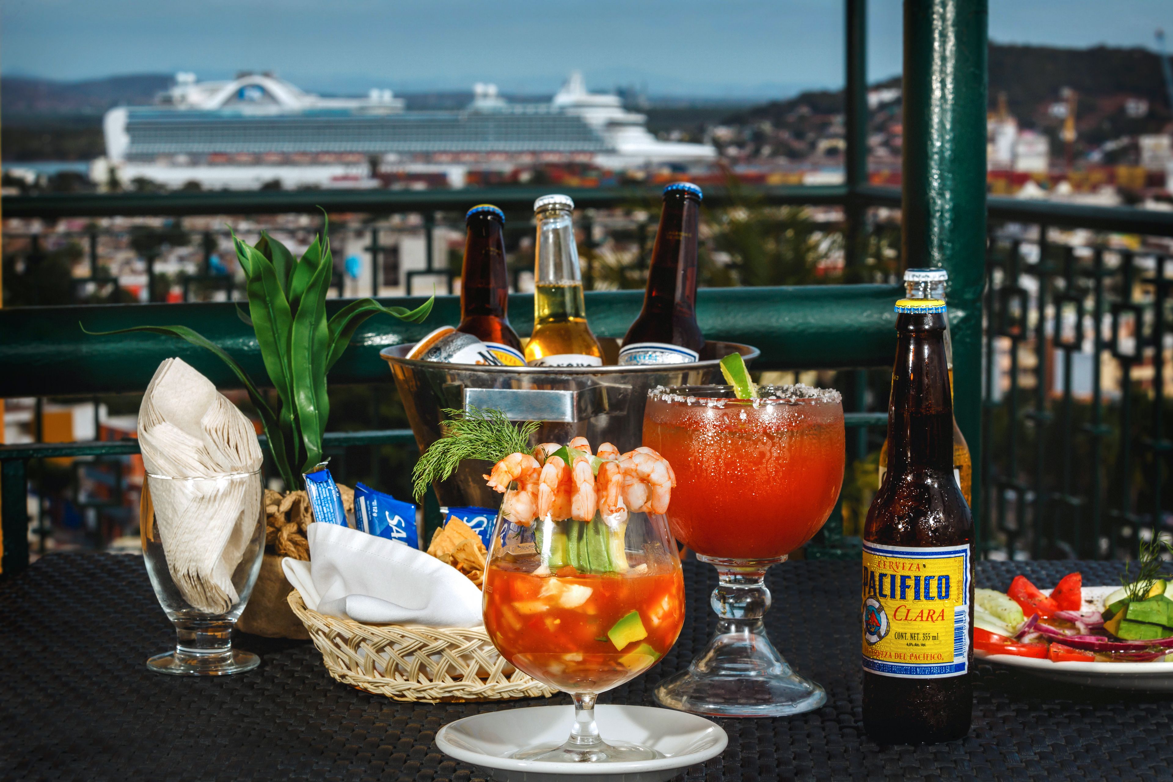 Een tafel met diverse drankjes, waaronder een cocktail met garnalen, een fles bier en een glas rode drank, met een mooi uitzicht op de achtergrond.