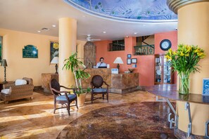 Um saguão de hotel com uma mesa de recepção, duas cadeiras de vime, um vaso de plantas, um relógio e um detalhe decorativo no teto.