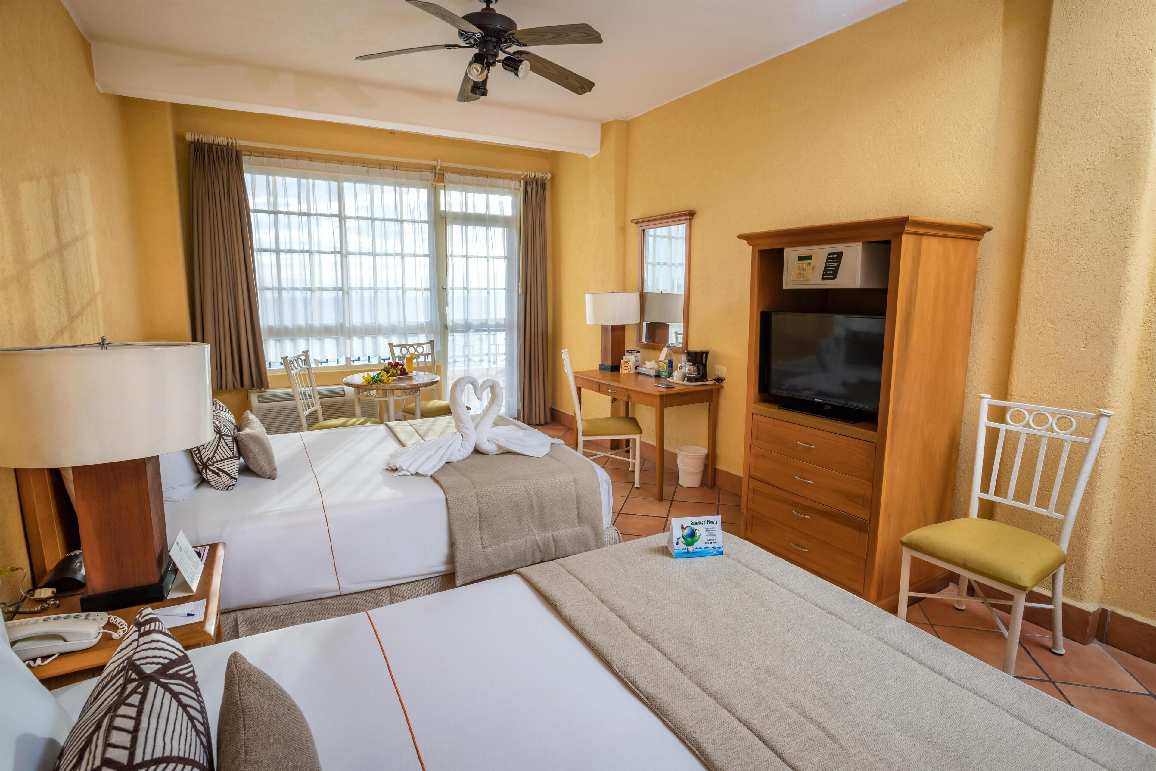 A hotel room with two beds, a desk with a television, a chair, and a dining area with a table and chairs.