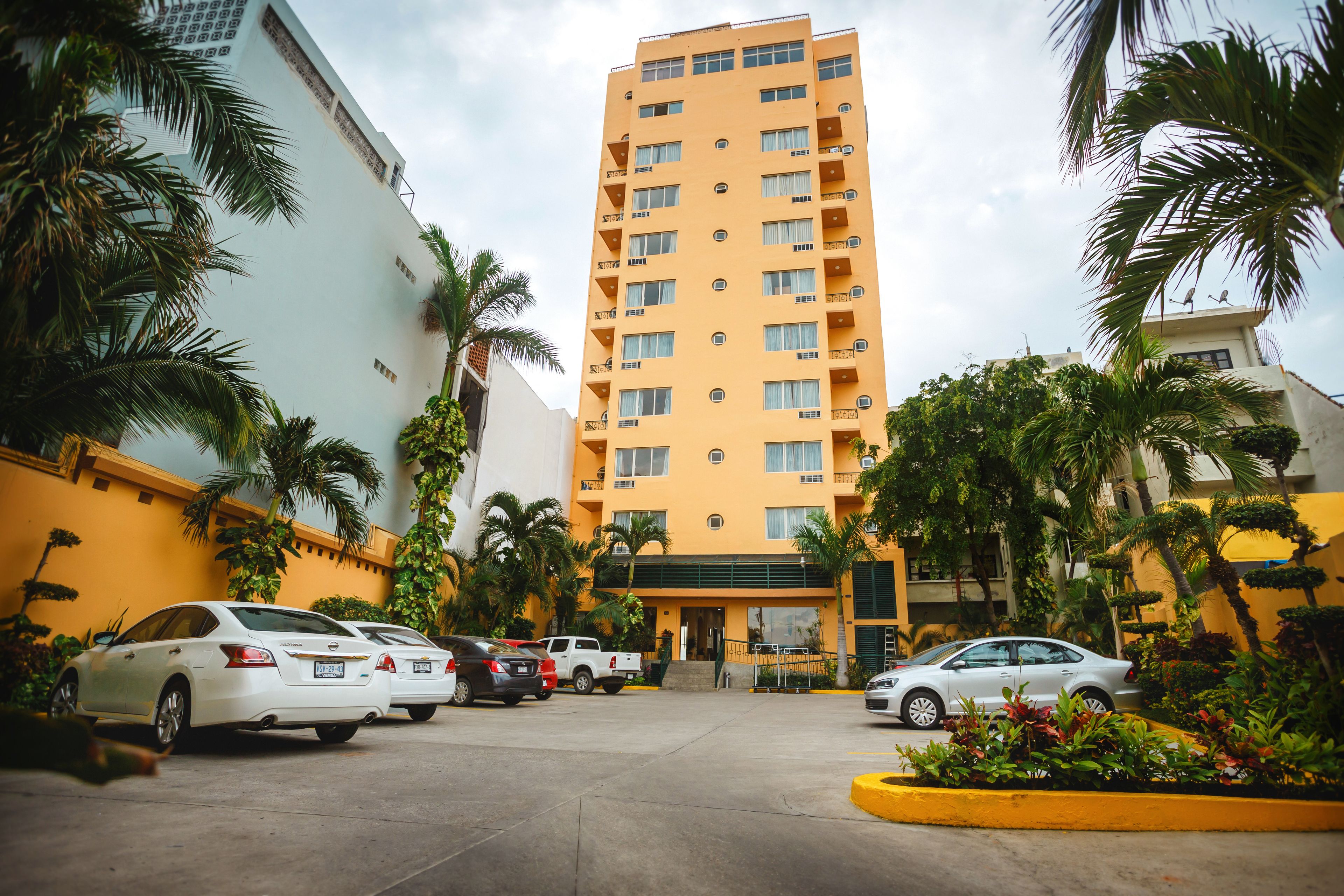Un hotel de varios pisos con fachada amarilla, coches estacionados y palmeras.