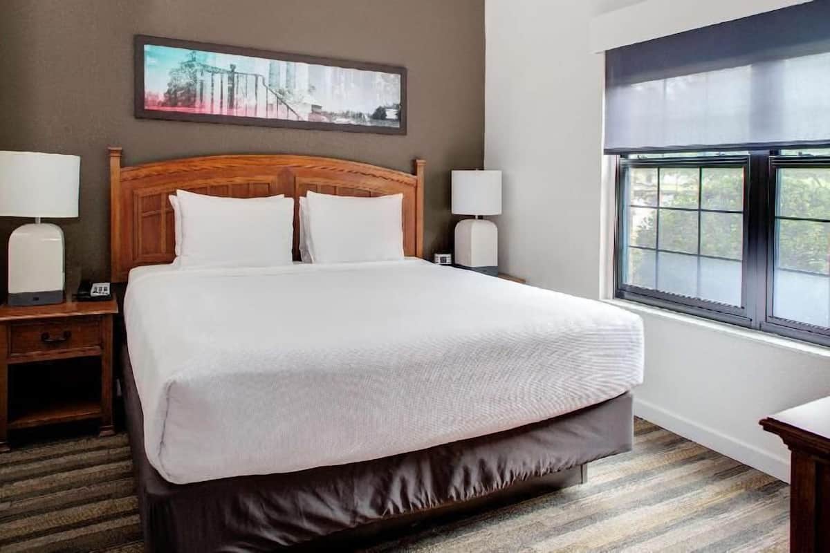 Habitación de hotel con una cama grande, cabecera de madera, dos mesitas de noche con lámparas, un cuadro en la pared y una ventana con vistas.