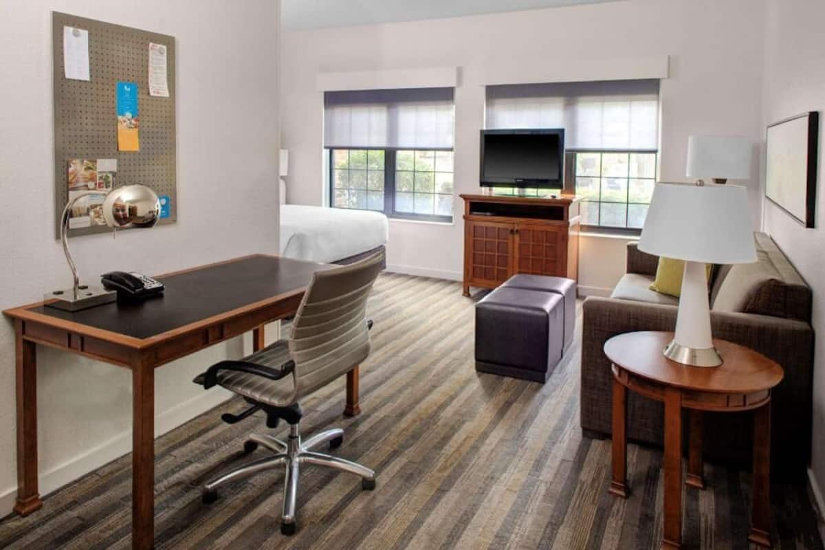 Habitación de hotel con cama, escritorio, silla, televisor, sofá y un tablón de anuncios.
