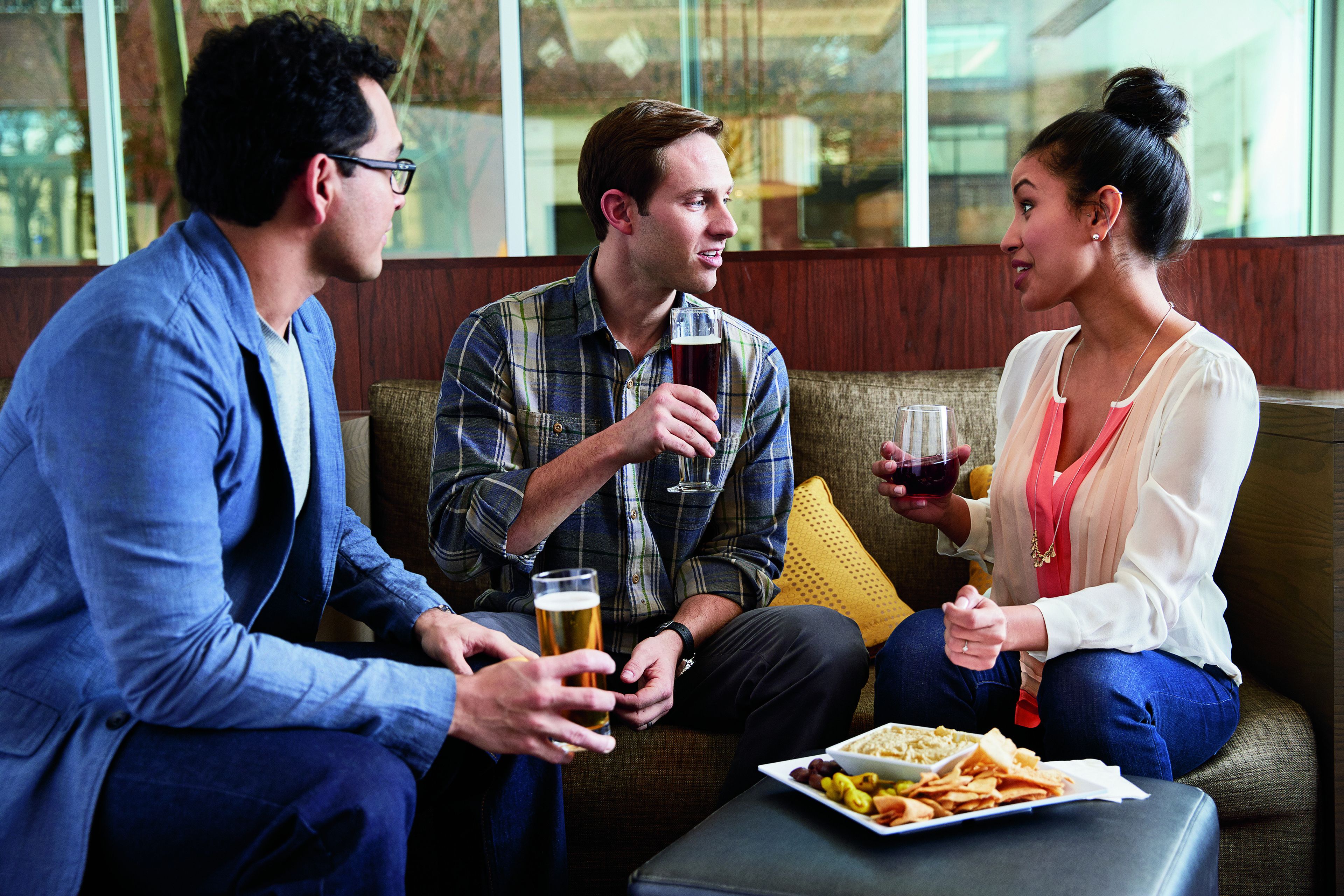Three people seated on a couch, holding glasses of beer, with a plate of snacks on a table in front of them.